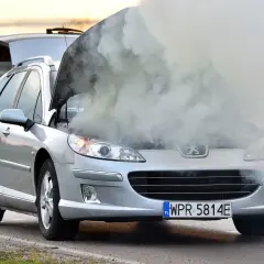 Srebrny Peugeot z otwartą maską i kłębami dymu. Rozbiegany diesel automat na poboczu drogi.